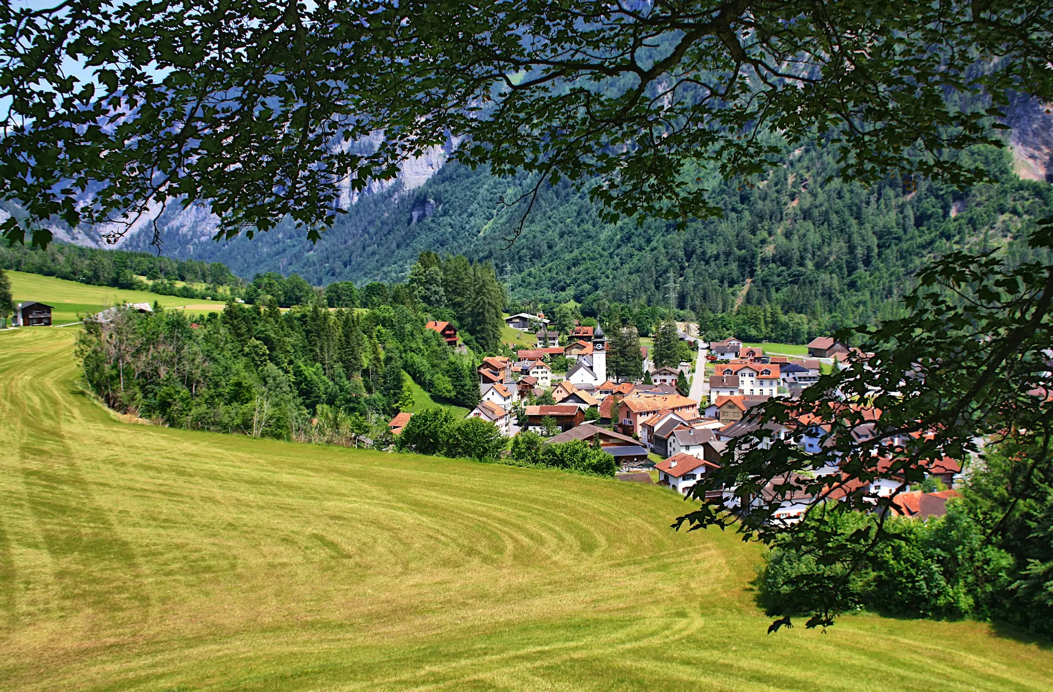 A picturesque village with a prominent white church steeple is situated in a verdant valley, framed by dark green tree…