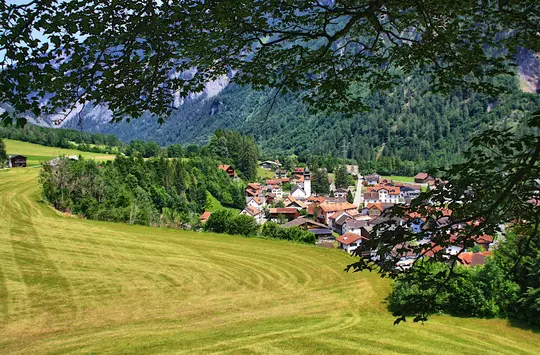 A picturesque village with a prominent white church steeple is situated in a verdant valley, framed by dark green tree…