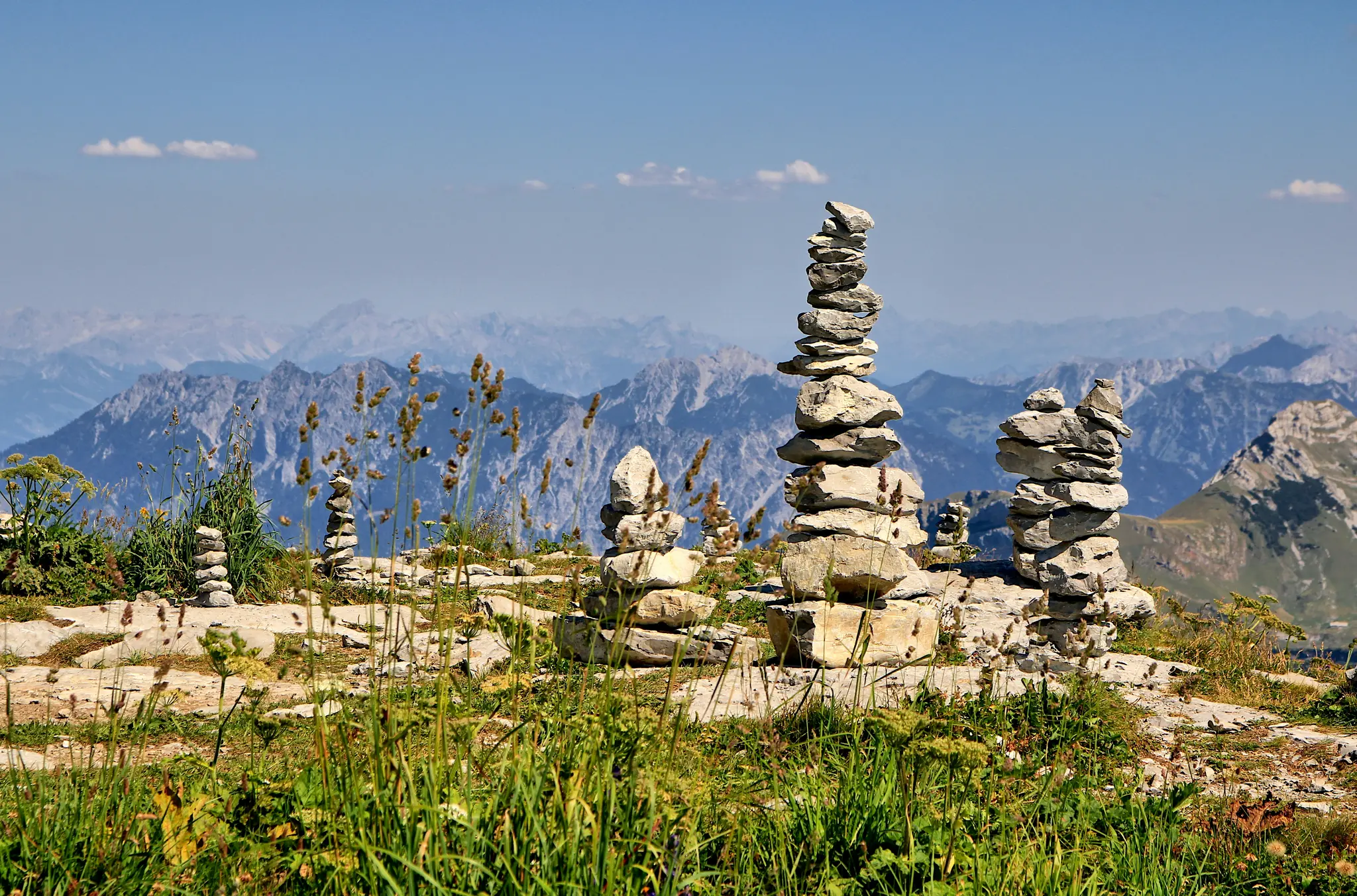 Several cairns, or stacks of rocks, stand on a rocky and grassy mountain ridge. In the background, layers of blue mountains…