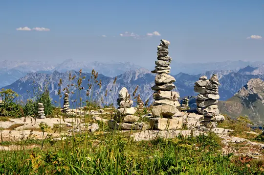 Several cairns, or stacks of rocks, stand on a rocky and grassy mountain ridge. In the background, layers of blue mountains…