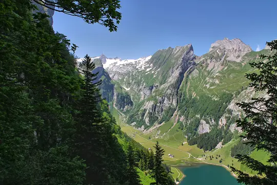 Panoraamanäkymä vehreästä vuoristolaaksosta, jossa on turkoosi järvi. Lumihuippuiset vuoret kohoavat taustalla sinisen