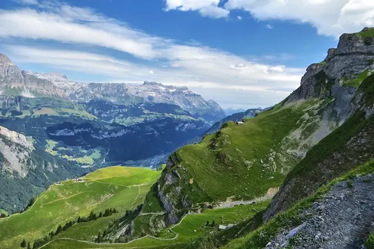 A wide landscape photo shows a vast mountain range with steep green slopes and rocky cliffs under a bright blue sky with…