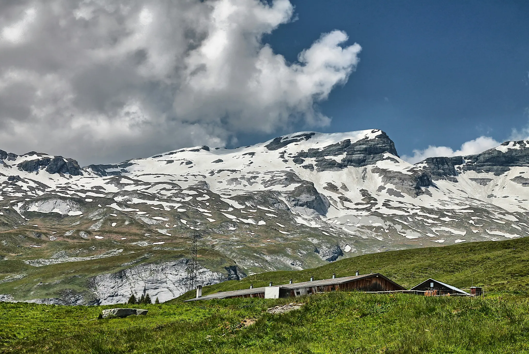 A wide landscape view of snow-capped mountains under a partly cloudy sky, with green grassy hills and a few wooden…