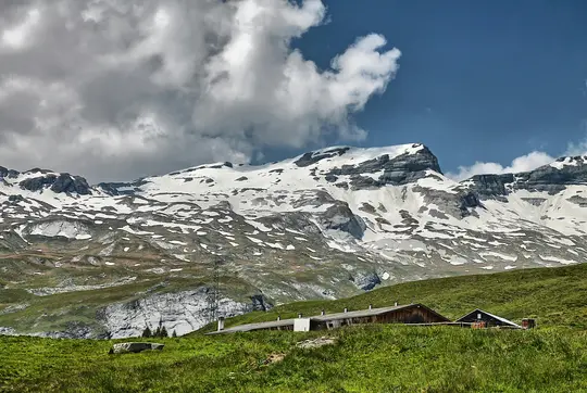 A wide landscape view of snow-capped mountains under a partly cloudy sky, with green grassy hills and a few wooden…