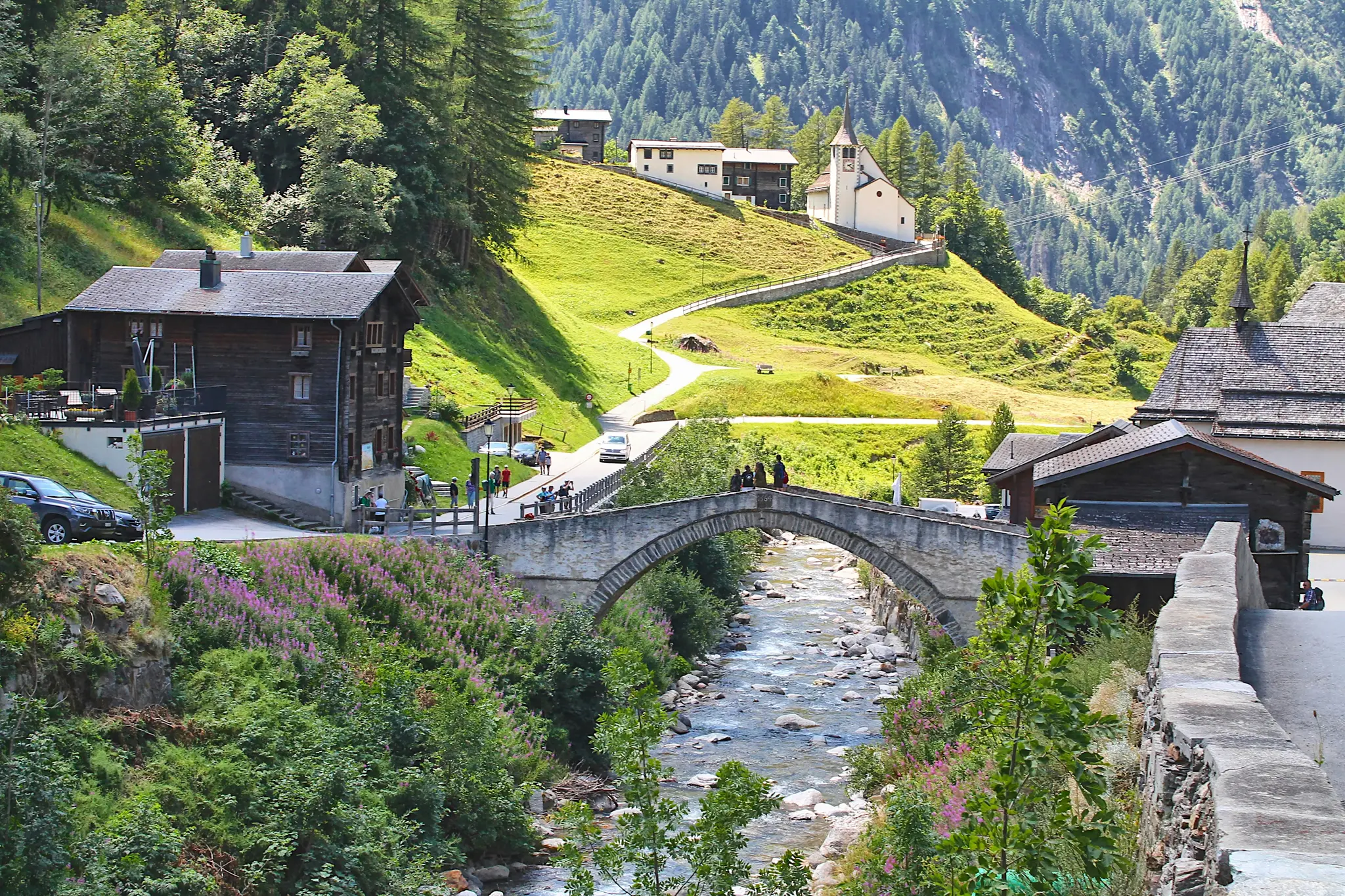 A picturesque mountain village with traditional wooden houses and a white church on a hillside. A river flows under a stone…