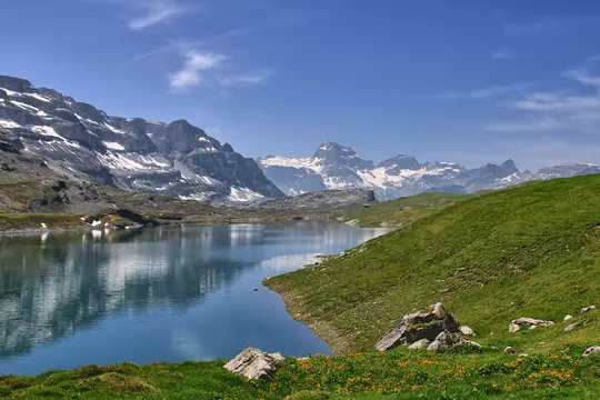 A wide landscape view of a calm blue lake reflecting snow-capped mountains under a clear blue sky with sparse clouds. Green…