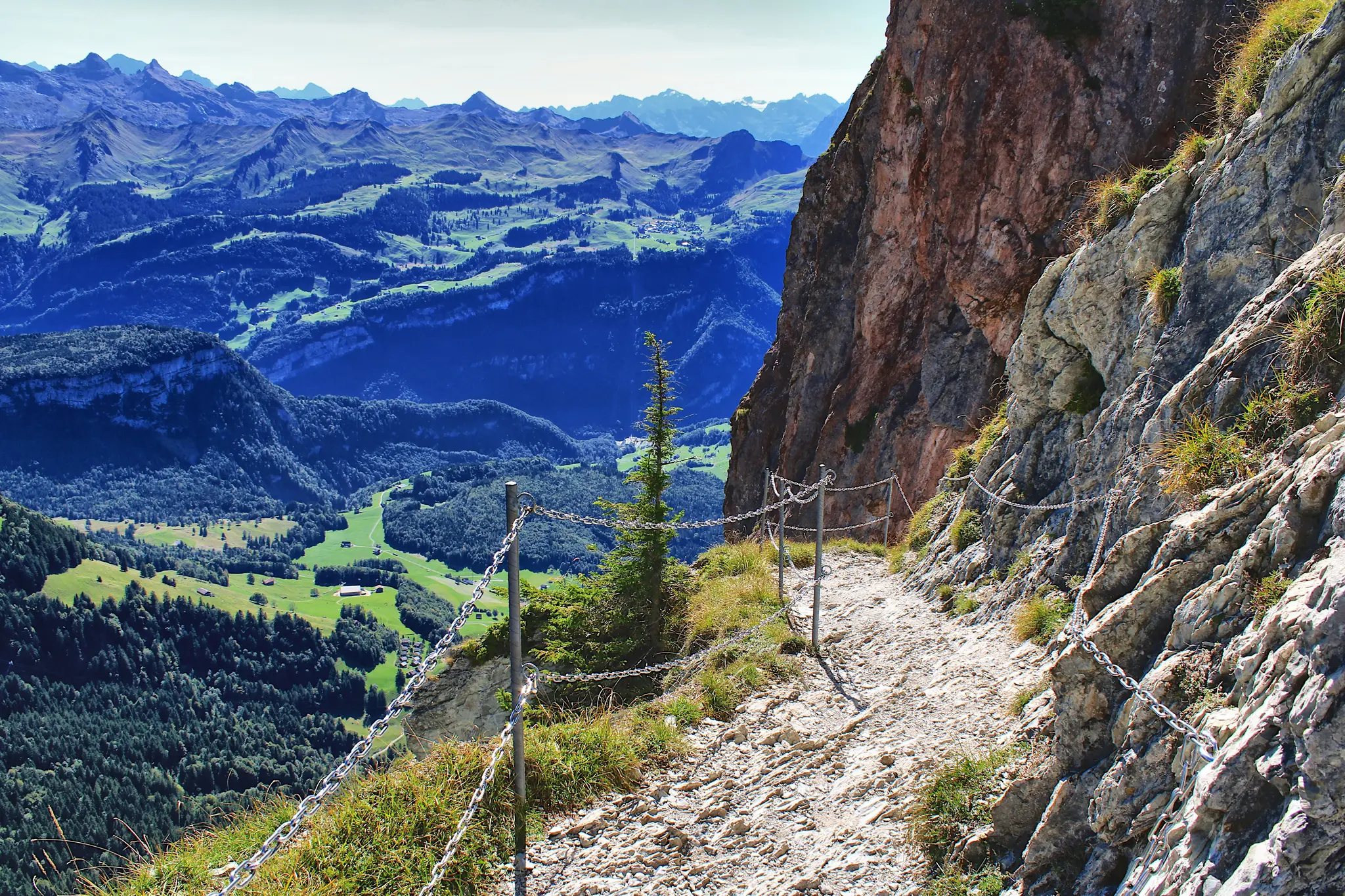 A narrow, rocky mountain path with a metal chain railing on the right, overlooking a deep valley with green fields and…