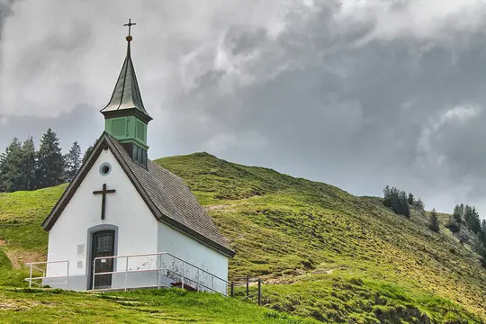 Et lille hvidt kapel med en grøn spir og en brun dør står på en levende grøn bjergskråning. Mørke skyer fylder himlen.