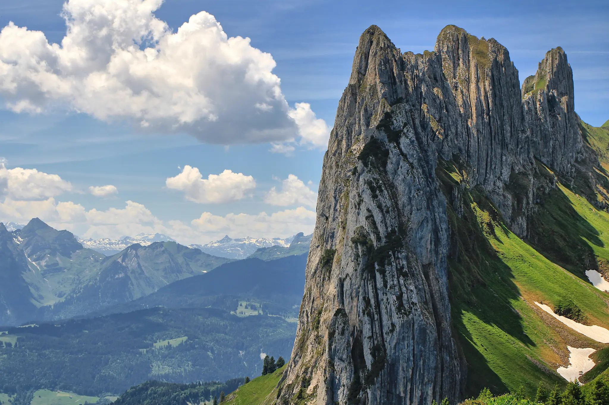 A grand landscape photo of a rugged, grey rocky mountain peak dominating the right side, with green grassy slopes and…