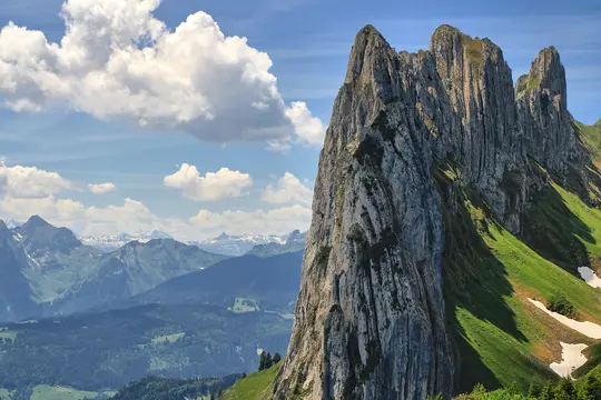 A grand landscape photo of a rugged, grey rocky mountain peak dominating the right side, with green grassy slopes and…