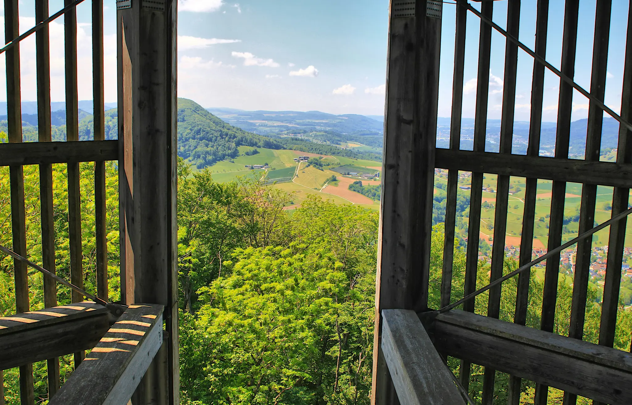 A view from a wooden lookout tower showing a wide valley with green forests, cultivated fields, and distant towns under a…