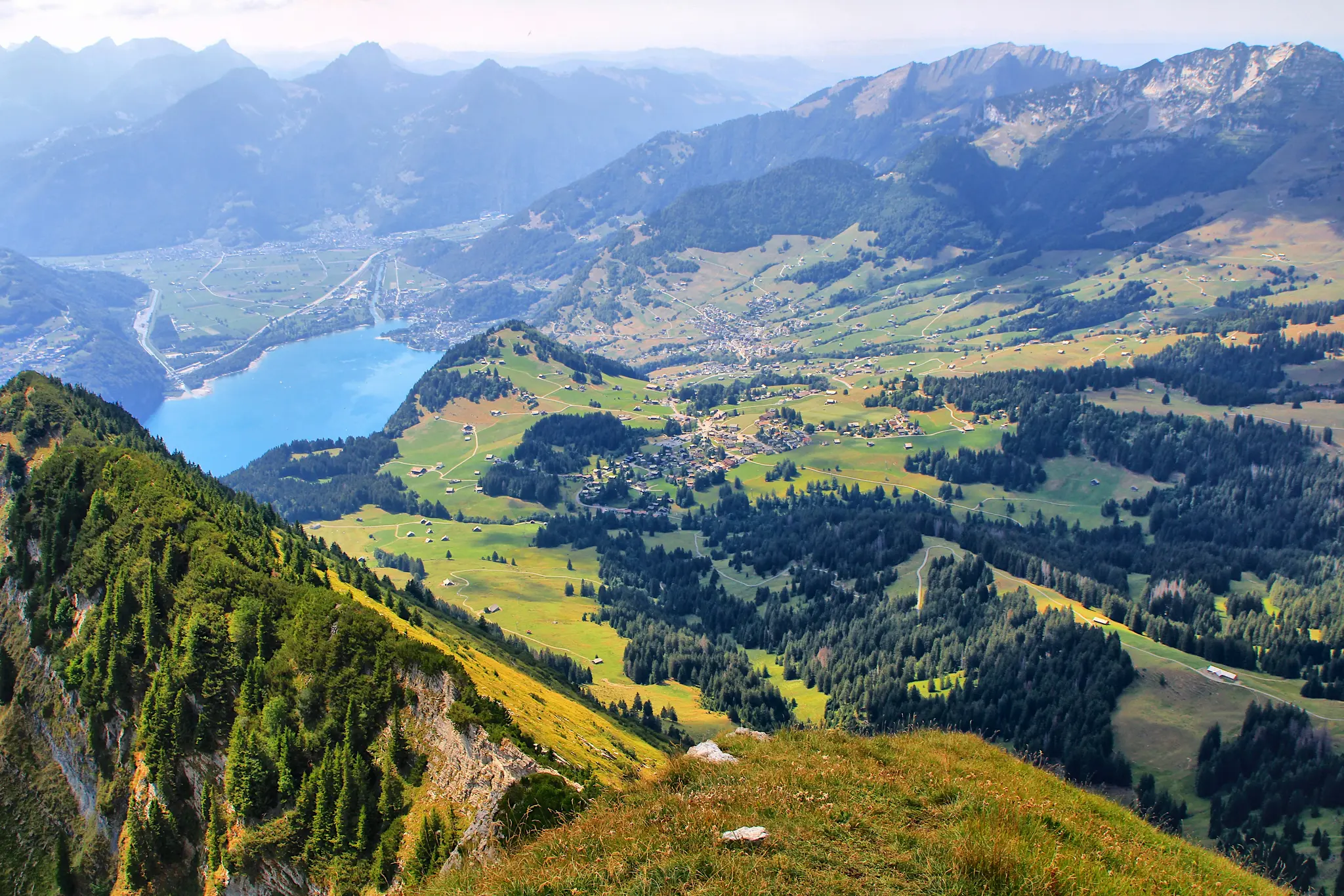 An aerial landscape photo shows a bright blue lake in a valley, surrounded by green mountains, forests, and small villages.