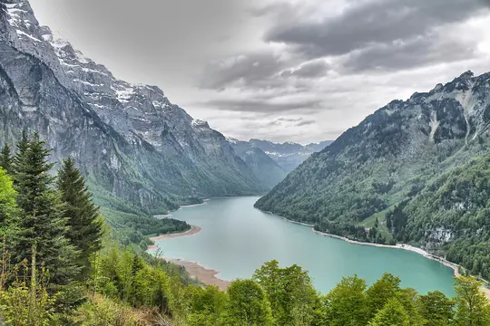 Panoraamanäkymä turkoosista järvestä, joka kiemurtelee laakson halki, reunustettuna jylhillä vuorilla, joissa on vihreitä