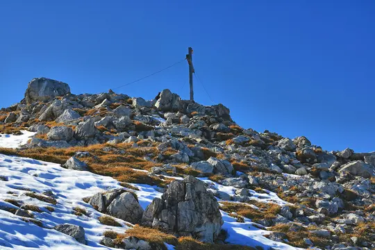 A clear blue sky dominates the upper half of the image, above a rocky mountain slope. Patches of white snow and dry brown…