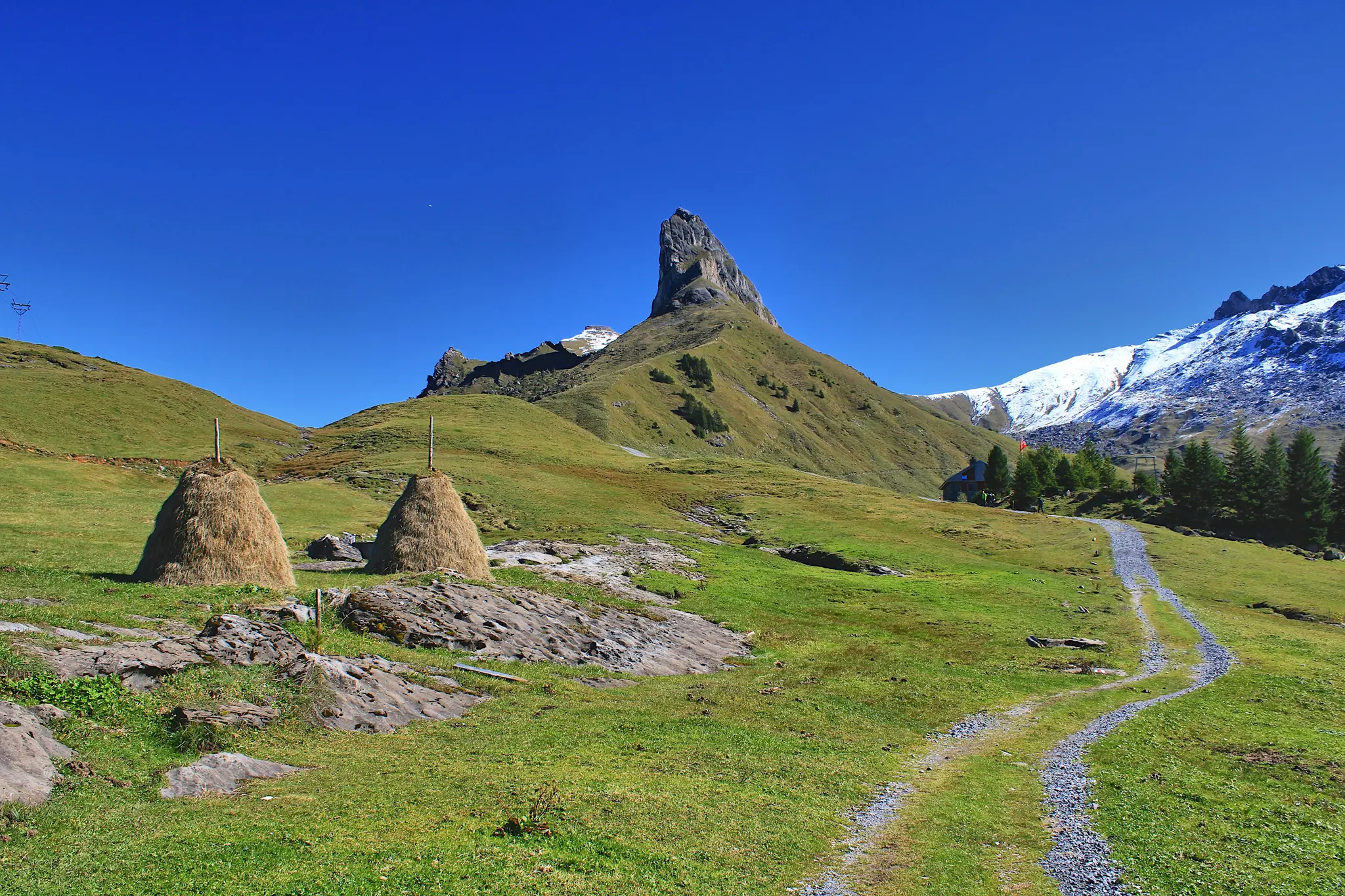 A wide landscape photo shows two conical haystacks in a green grassy field with scattered rocks. A gravel path winds…