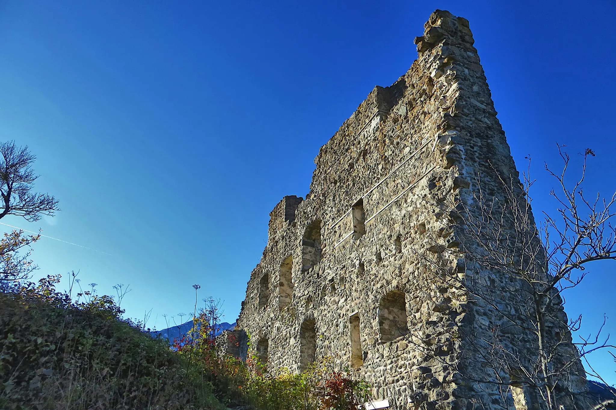 A low-angle view of a tall, crumbling stone wall, part of an ancient ruin, under a bright, clear blue sky. Bare tree…