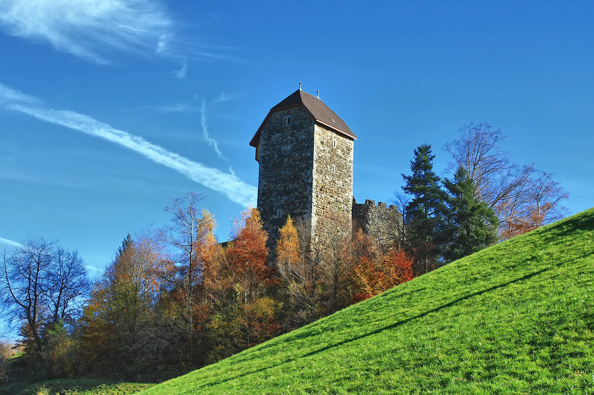 A tall, square stone castle tower with a dark brown roof stands against a bright blue sky with wispy clouds. Below it, a…