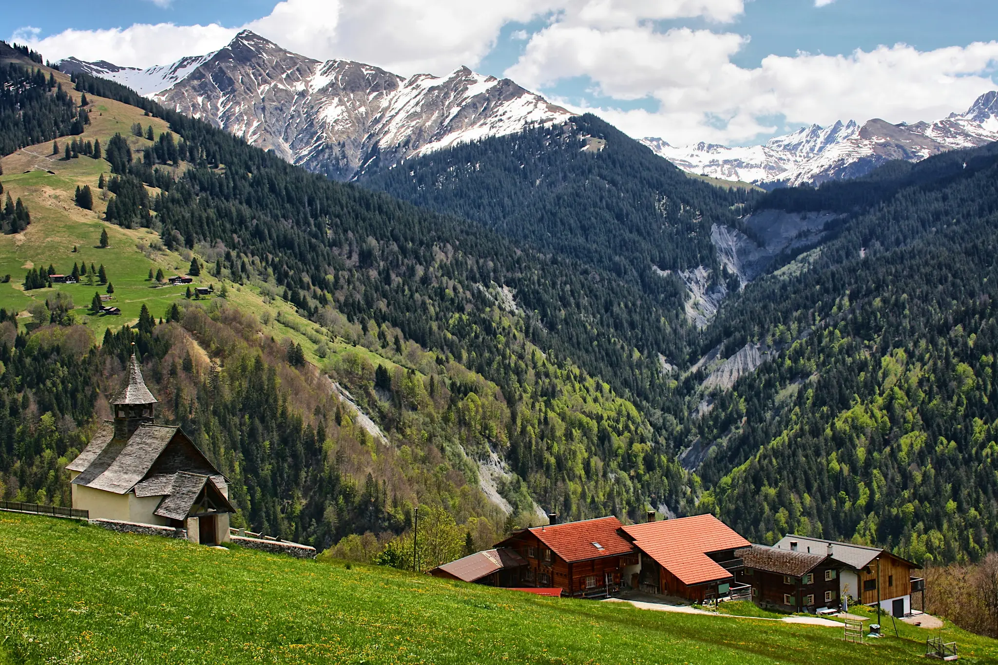 A scenic view of an alpine valley featuring a small church and several houses on a vibrant green hillside. Snow-capped…