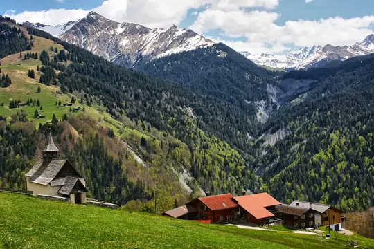 A scenic view of an alpine valley featuring a small church and several houses on a vibrant green hillside. Snow-capped…