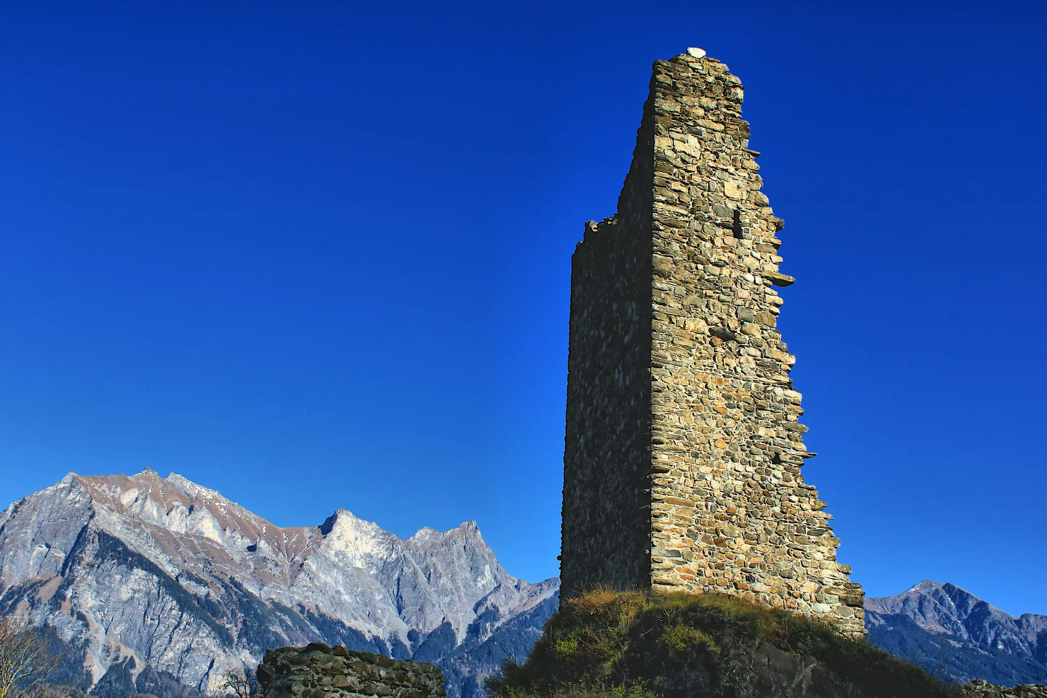 A tall, crumbling stone tower stands prominently on the right against a vibrant blue sky, with rugged mountains in the…