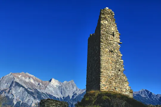 A tall, crumbling stone tower stands prominently on the right against a vibrant blue sky, with rugged mountains in the…