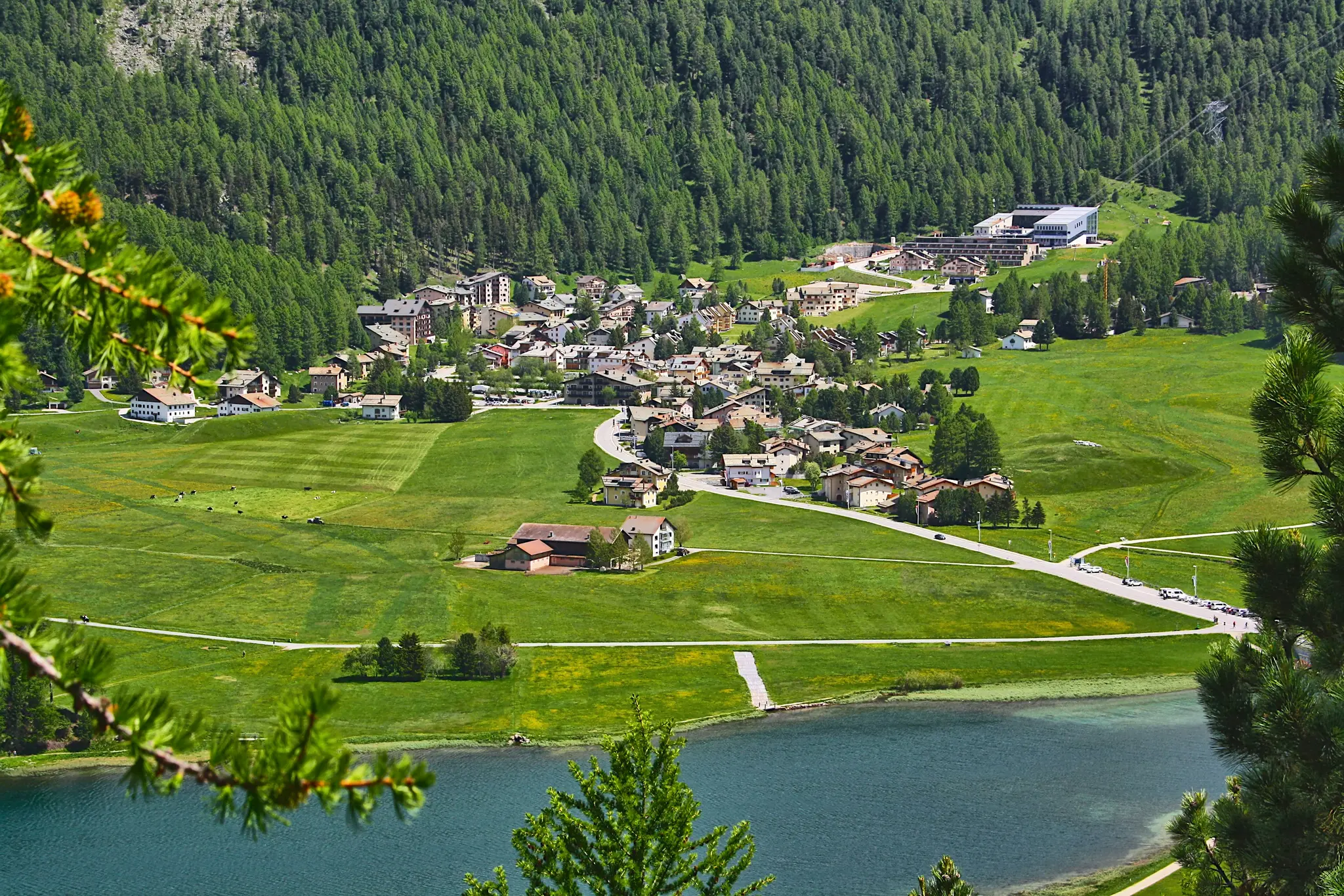 An aerial view shows a village with many buildings and winding roads in a lush green valley next to a blue lake. A dense…