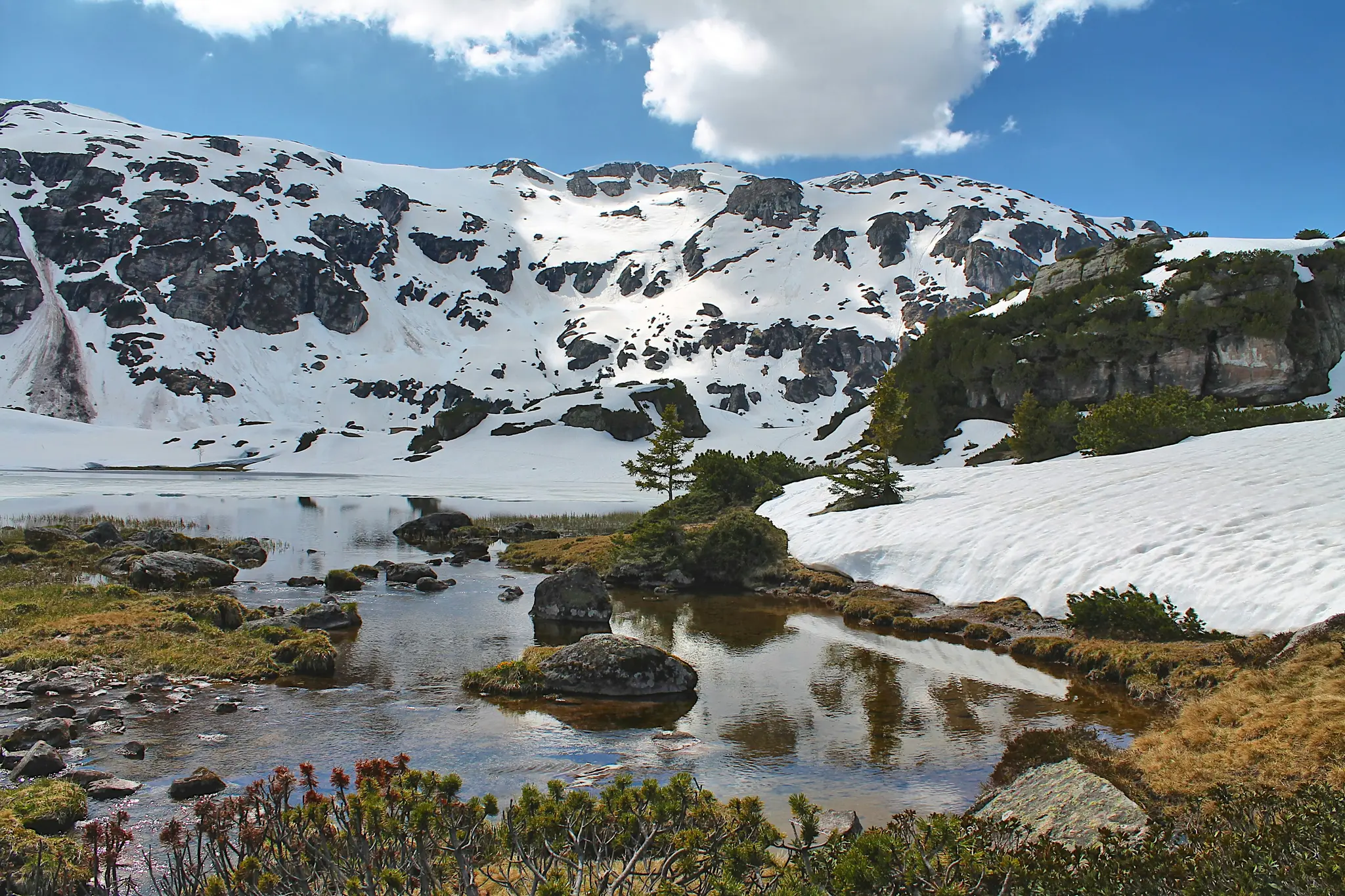 A wide landscape view of snow-covered mountains reflecting in a partially frozen lake under a bright blue sky with white…