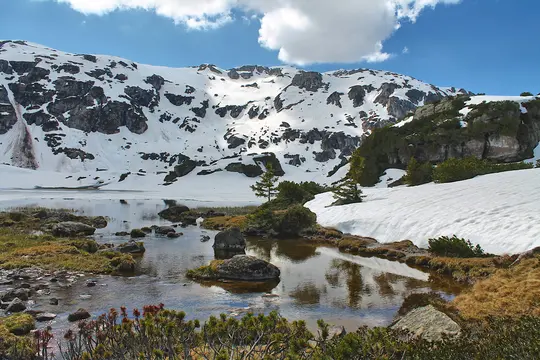 A wide landscape view of snow-covered mountains reflecting in a partially frozen lake under a bright blue sky with white…