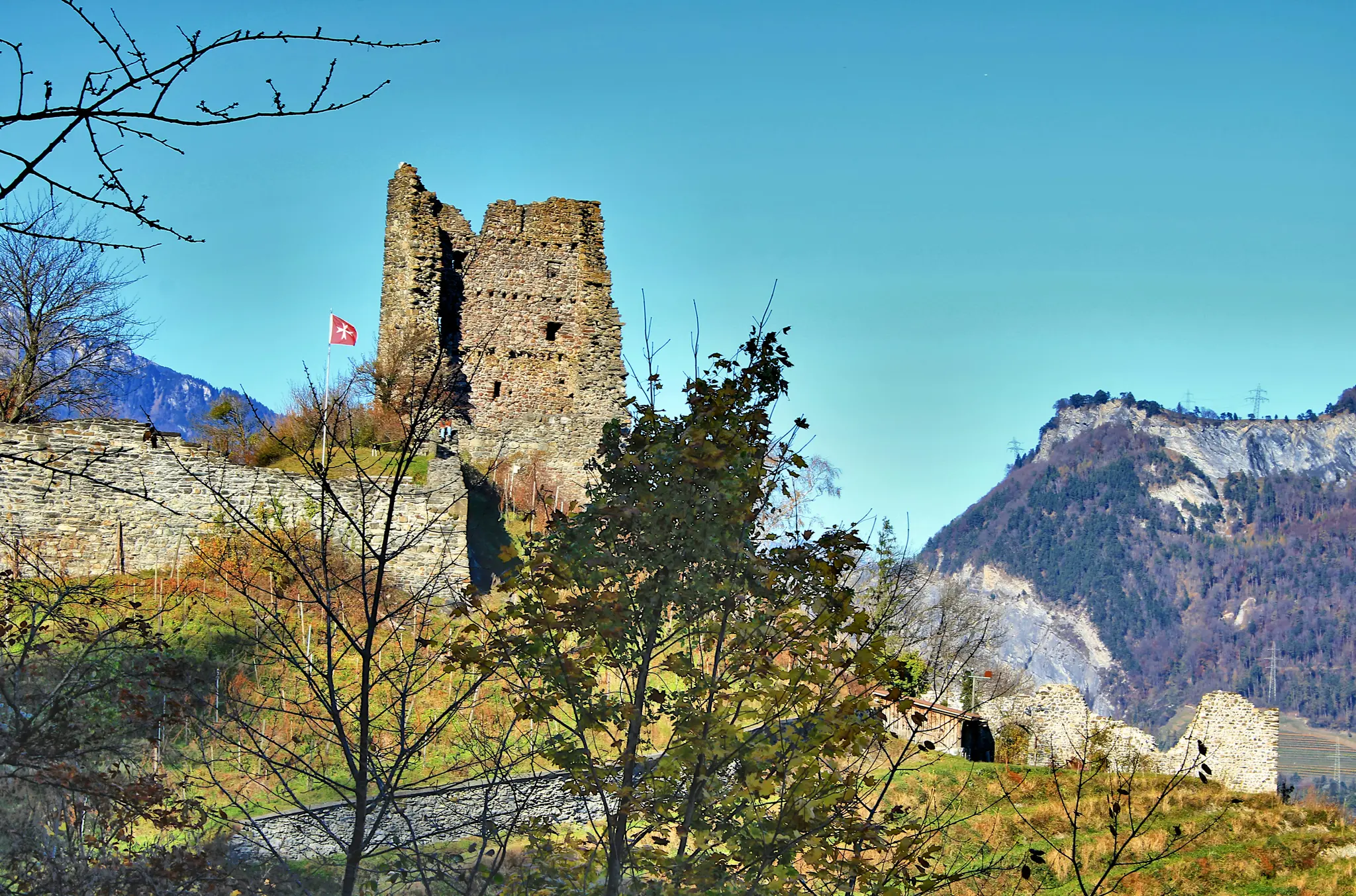 A wide landscape view of ancient stone castle ruins on a grassy hill, with a red flag flying. Mountains rise in the…