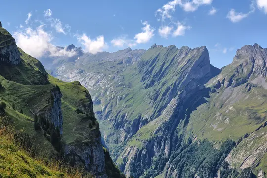 A panoramic view of a vast mountain range with lush green slopes, jagged grey peaks, and a bright blue sky with white clouds.