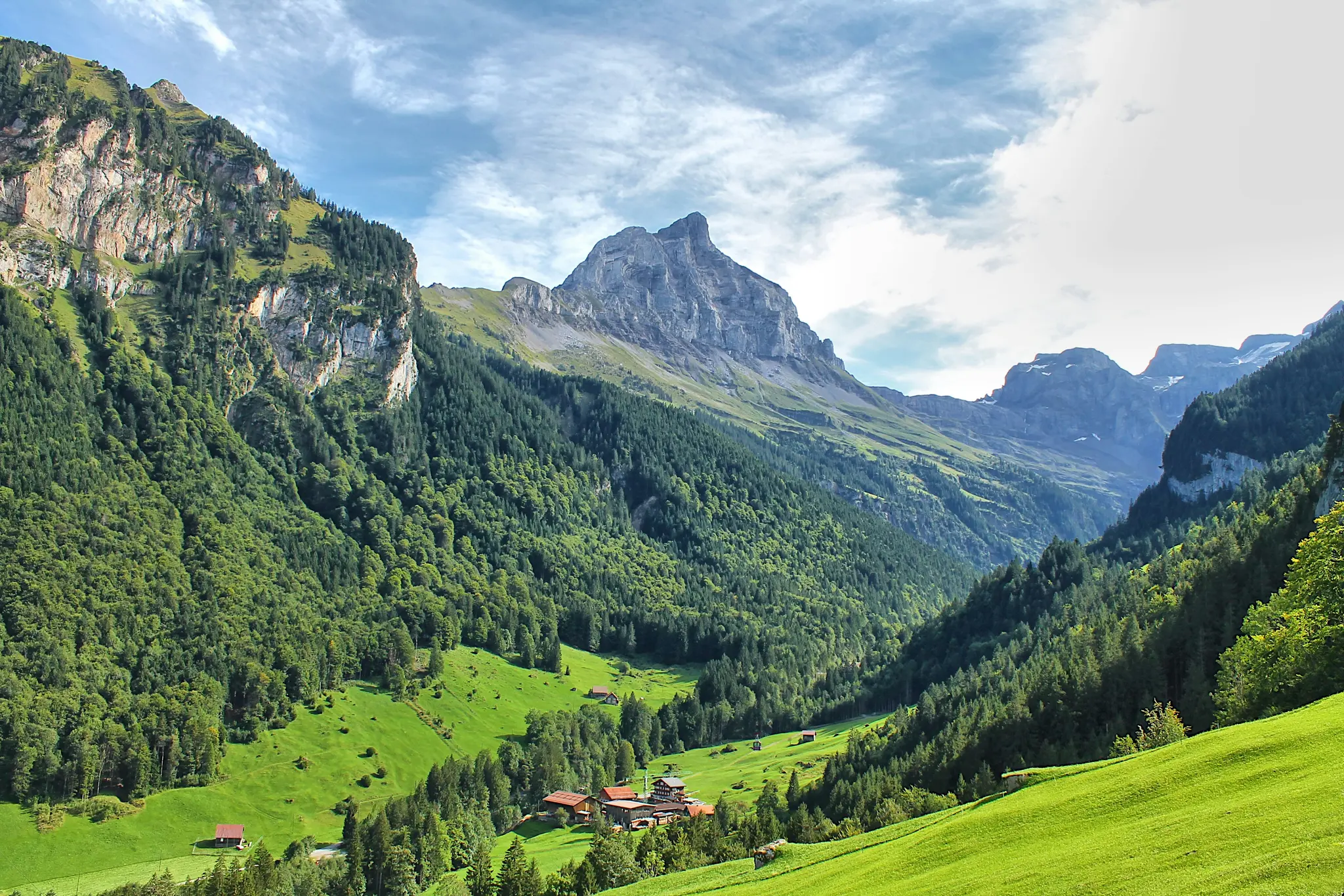 A panoramic view of a lush green valley with a small cluster of buildings, surrounded by dense forests and tall, rocky…