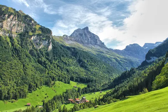 A panoramic view of a lush green valley with a small cluster of buildings, surrounded by dense forests and tall, rocky…