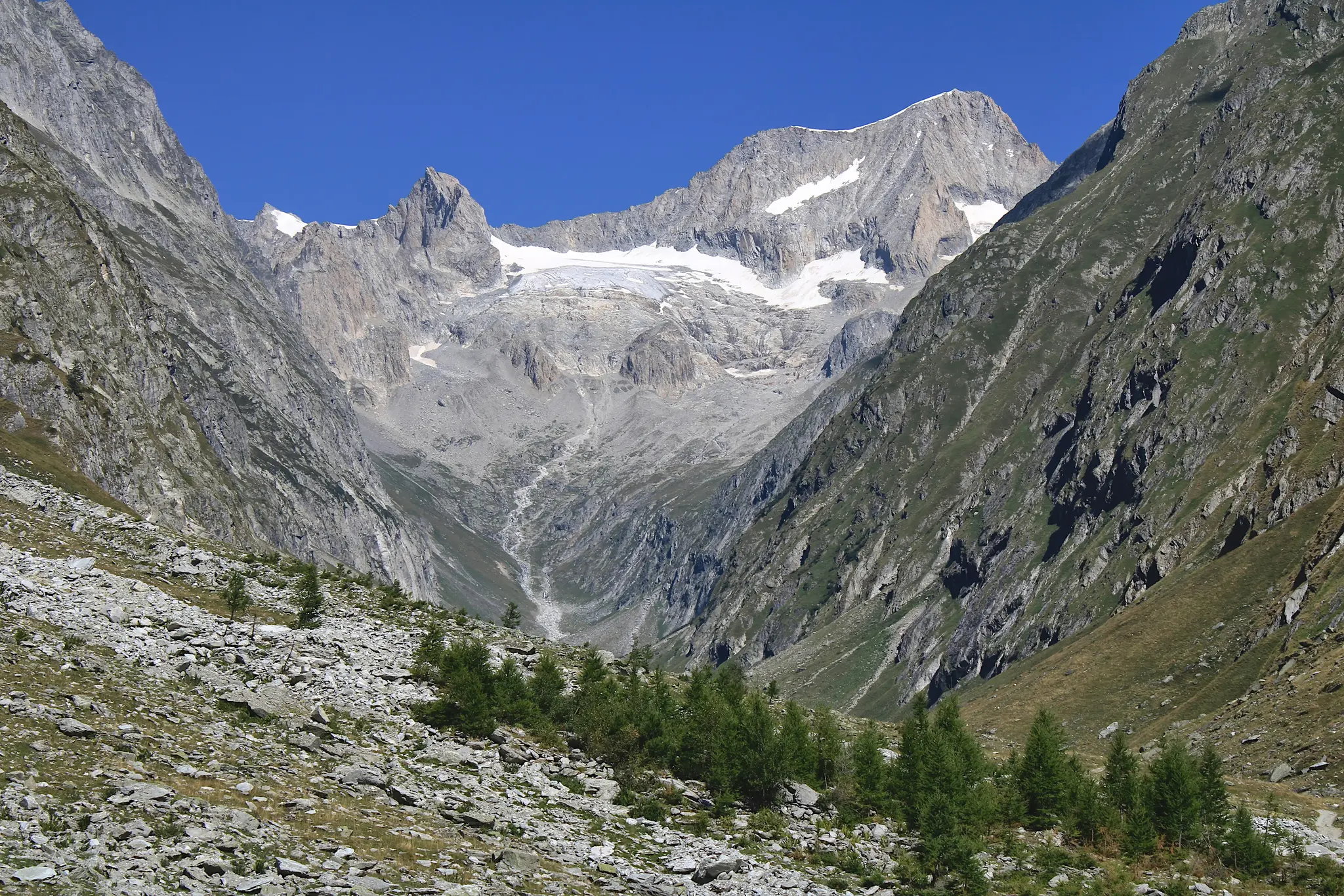 A wide landscape photo of a mountain valley. Rocky, grey slopes rise on either side, meeting a clear blue sky above. In the…