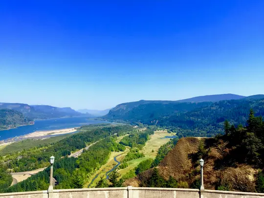 A panoramic view of a wide river flowing through a green valley with dense forests and distant mountains under a bright…