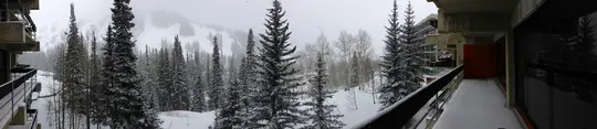 Široký panoramatický snímek zobrazuje zasněženou horskou krajinu s četnými jehličnany. Balkony budovy rámují…