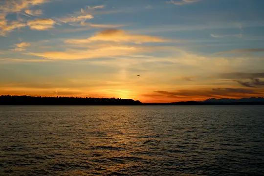 Živý západ slunce nad klidnou vodní hladinou s tmavou siluetou stromů a vzdálenými horami.