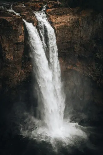 Une cascade haute et puissante plonge le long d'une falaise rocheuse sombre et texturée. L'eau blanche se brise dans une