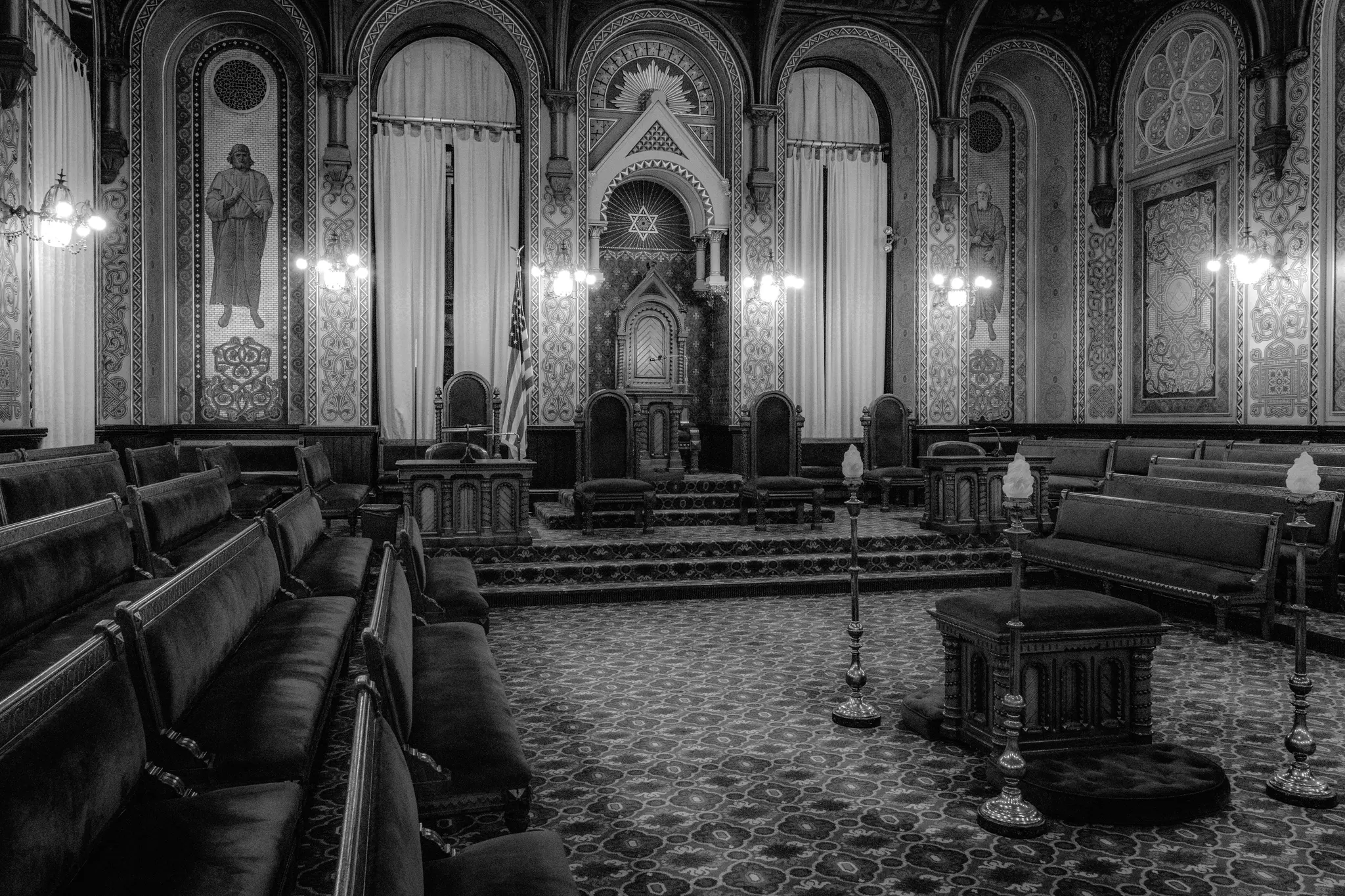 Une photo en noir et blanc d'une salle vide et ornée avec des rangées de bancs sombres faisant face à une estrade centrale