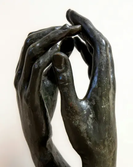 A close-up, vertical shot of a dark bronze sculpture depicting two hands with fingers intertwined, set against a plain…