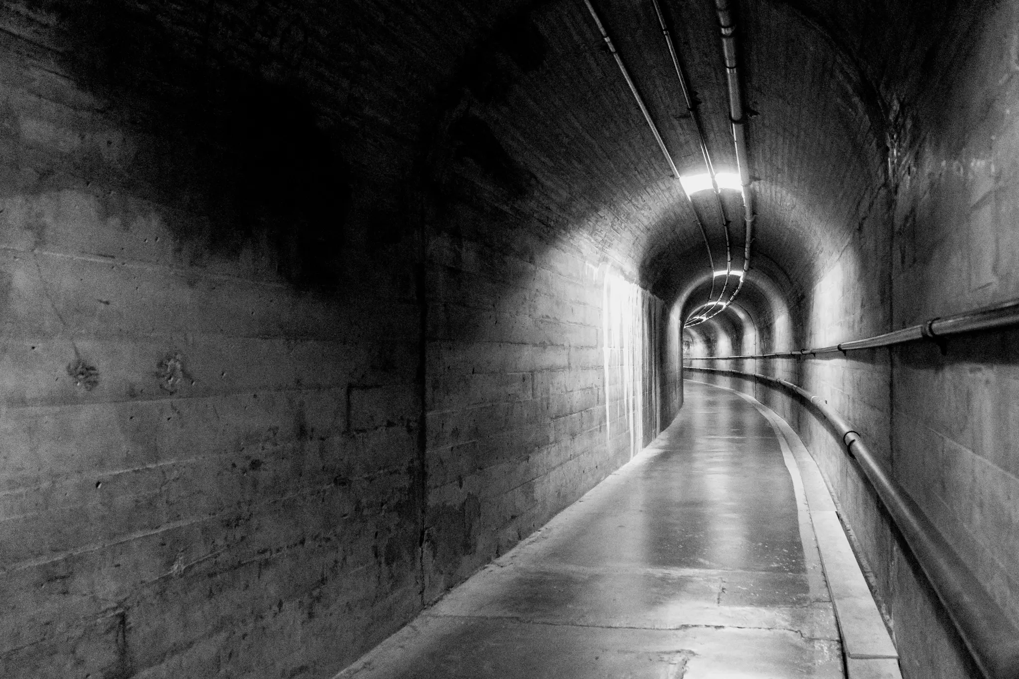 Čiernobiela fotografia dlhej, zakrivenej betónovej chodby. Stropné svetlá osvetľujú cestu a potrubia vedú pozdĺž…