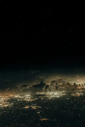 An aerial view of a sprawling city at night, illuminated by countless warm yellow lights, with dark clouds scattered above…