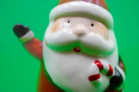 Detail keramickej figúrky Santa Clausa s bielym fúzom, červenou čiapkou a lízatkom na jasne zelenom pozadí.