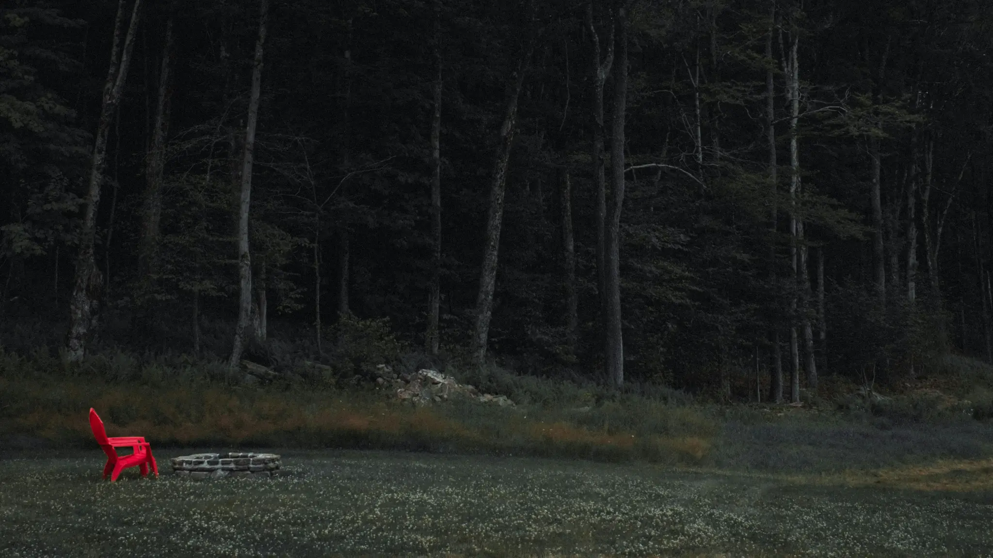 Živo červené kreslo Adirondack sedí vedľa kamenného ohniska na trávnatej čistinke, za ním tmavý, hustý les.