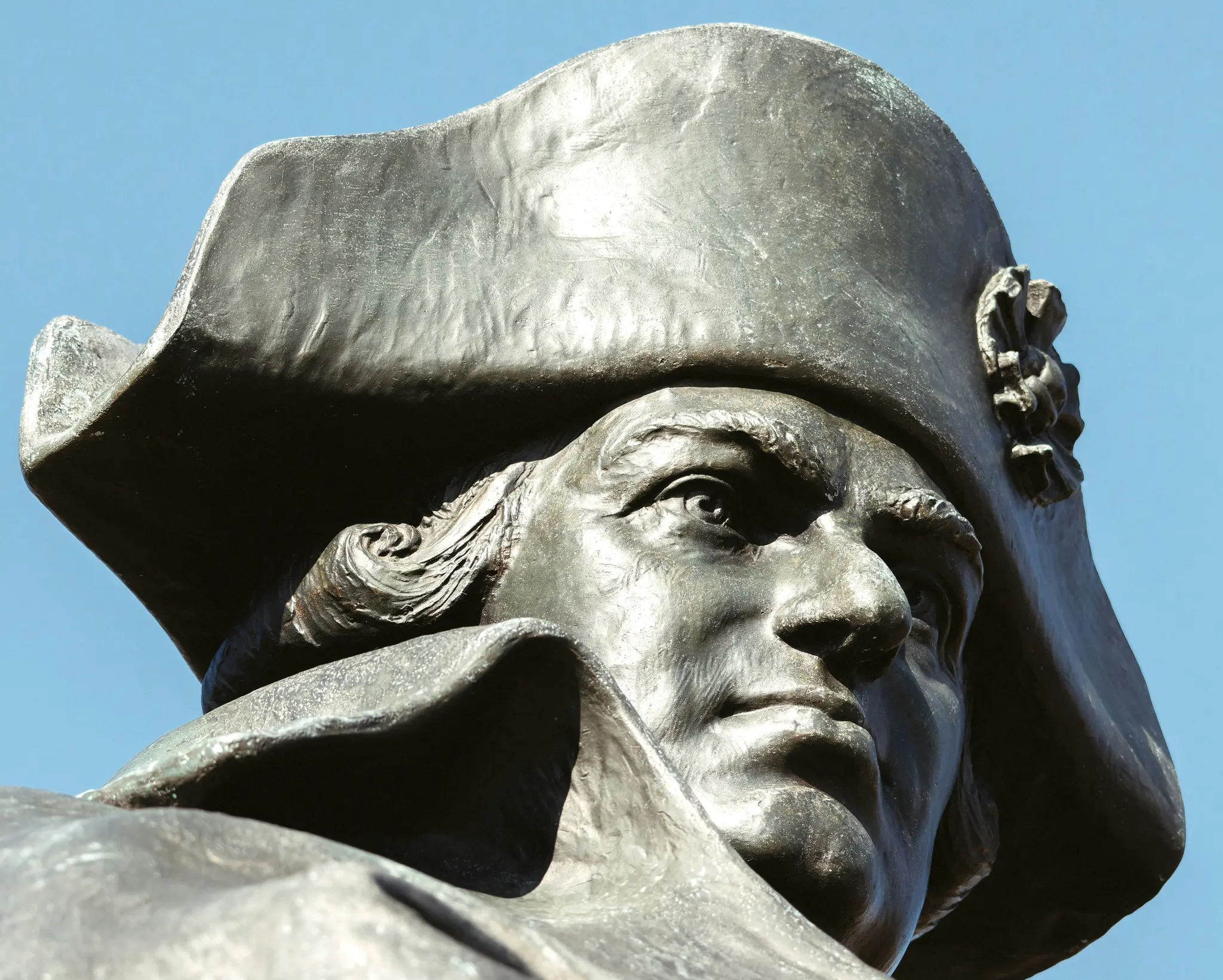 Gros plan en contre-plongée d'une statue en bronze d'un homme au bicorne, regardant vers la droite. Le ciel est d'un bleu
