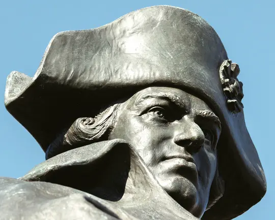 A low-angle, close-up shot of a bronze statue of a man in a bicorne hat, looking to the right. The sky is clear blue.
