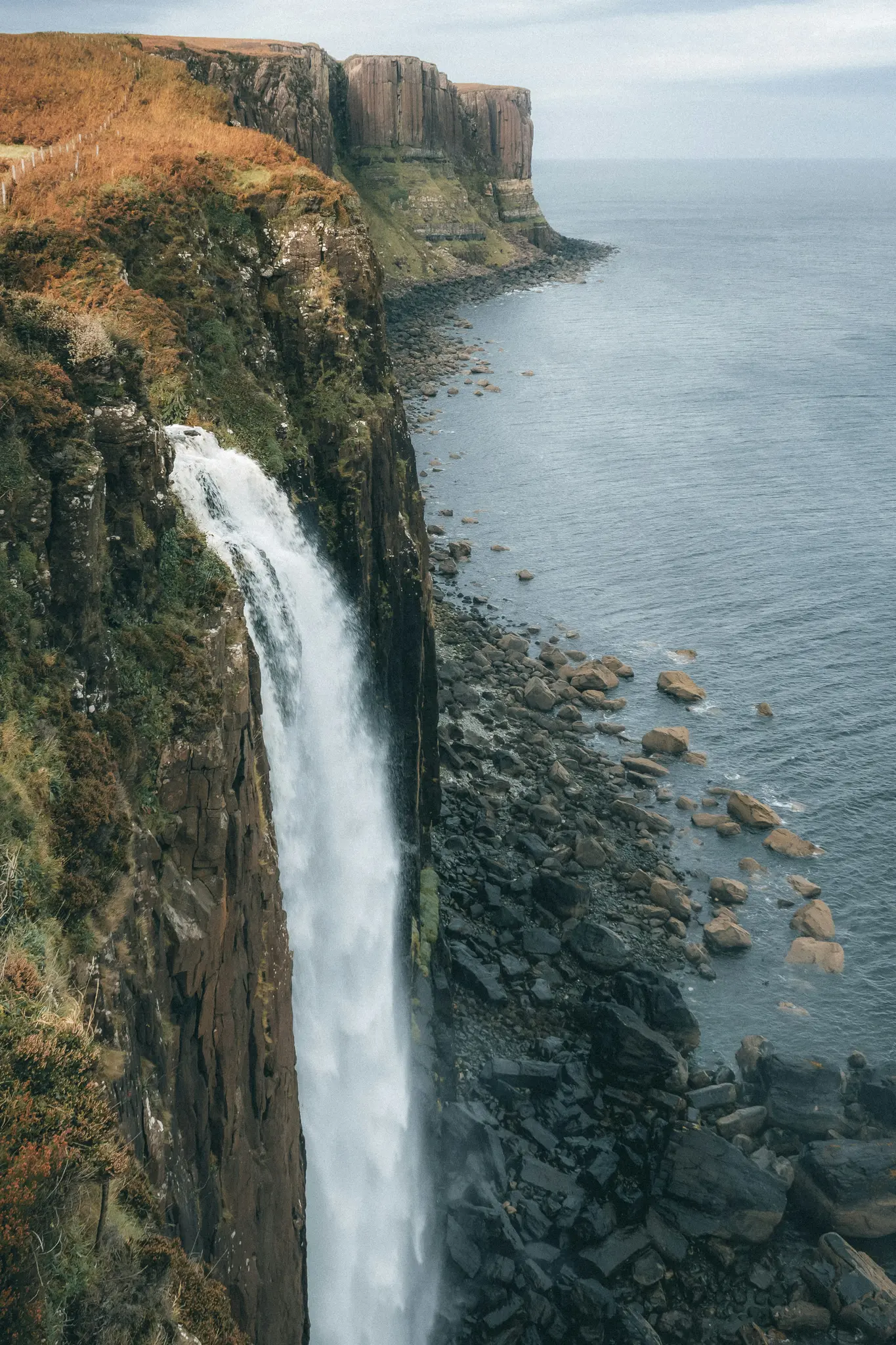 Une puissante cascade tombe d'une falaise haute et sombre dans la mer bleu foncé. Le bord de la falaise est couvert de