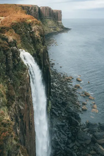 A powerful waterfall plunges from a tall, dark cliff into the dark blue sea. The cliff edge is covered in brown and green…