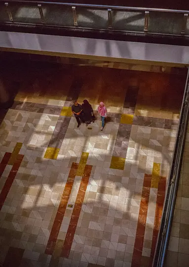 Záber zhora ukazuje troch ľudí na dláždenej podlahe s geometrickými vzormi v odtieňoch béžovej, hnedej a žltej. Silné…