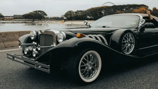 En sort klassisk bil med kromdetaljer og hvidvæggede dæk er parkeret på en asfaltvej. En hånd rækker ud fra…
