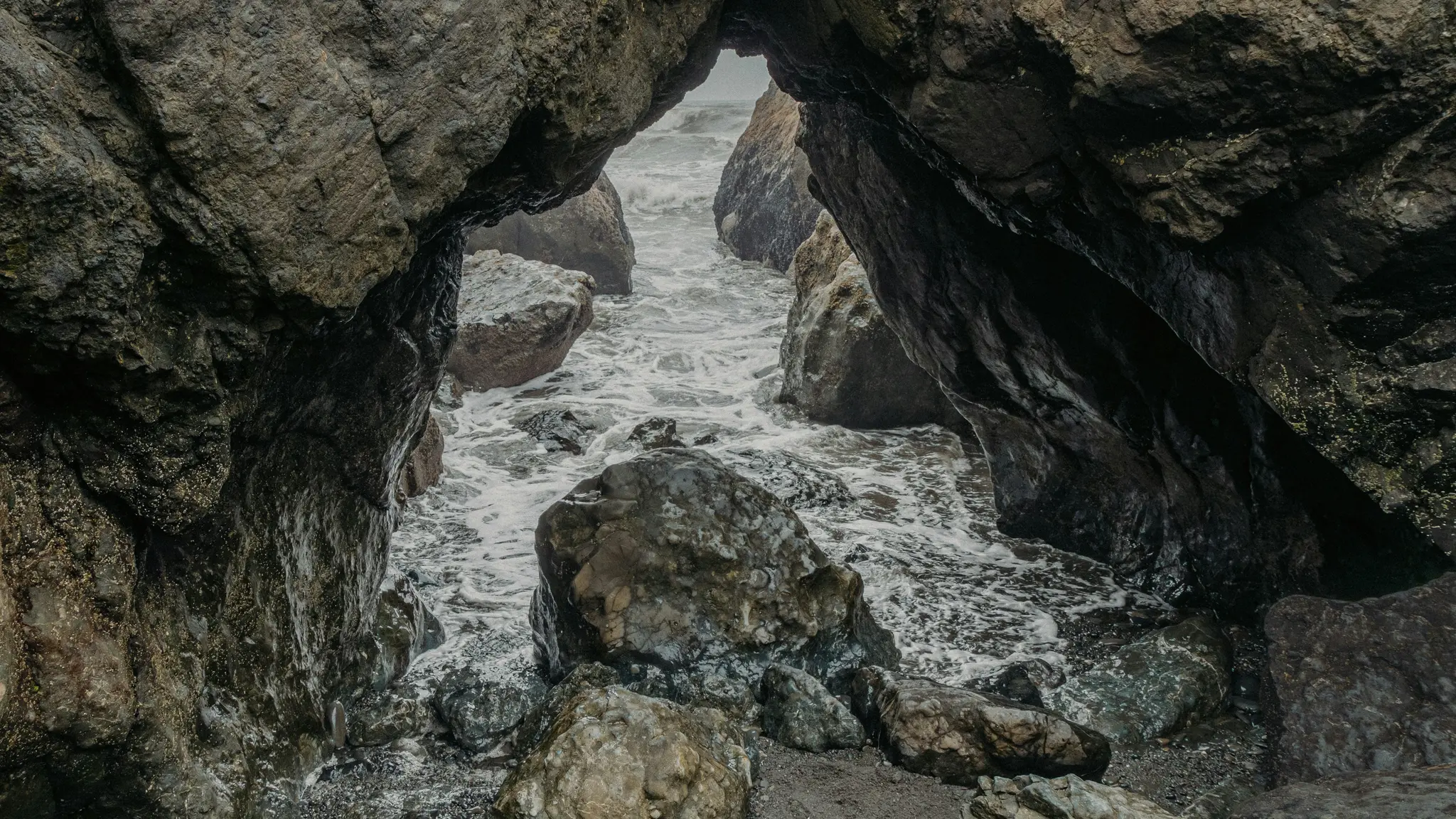 Une vue à travers une arche rocheuse sombre et accidentée sur l'océan au-delà. Des vagues écumeuses se brisent sur des